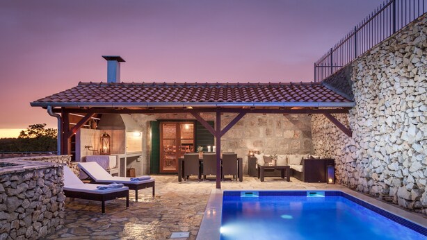 Un espacio al aire libre con paredes de piedra, una piscina, sillones de descanso y un porche cubierto.