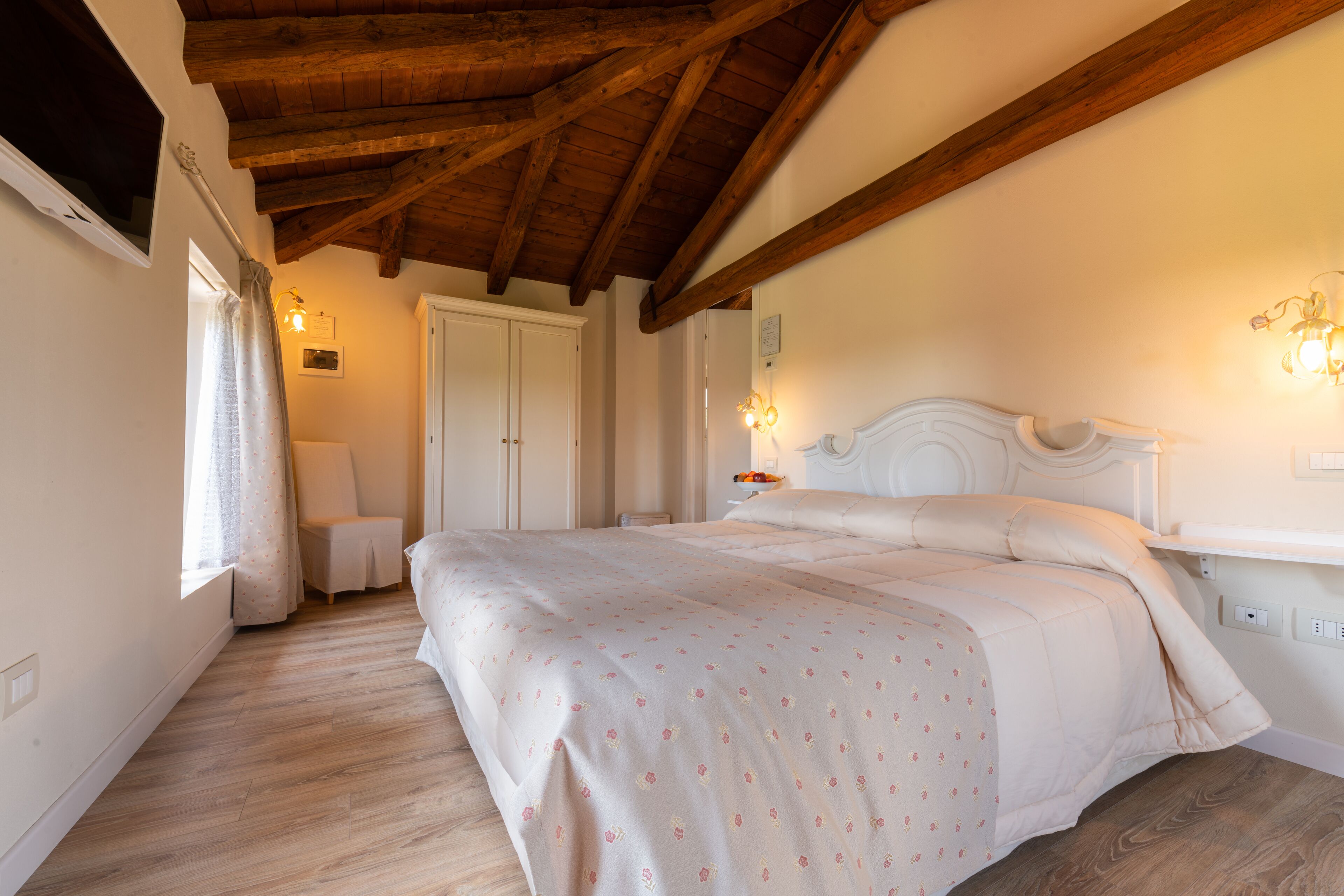 A bedroom with a wooden ceiling, a bed with a floral bedspread, a nightstand with a lamp, and a window with curtains.