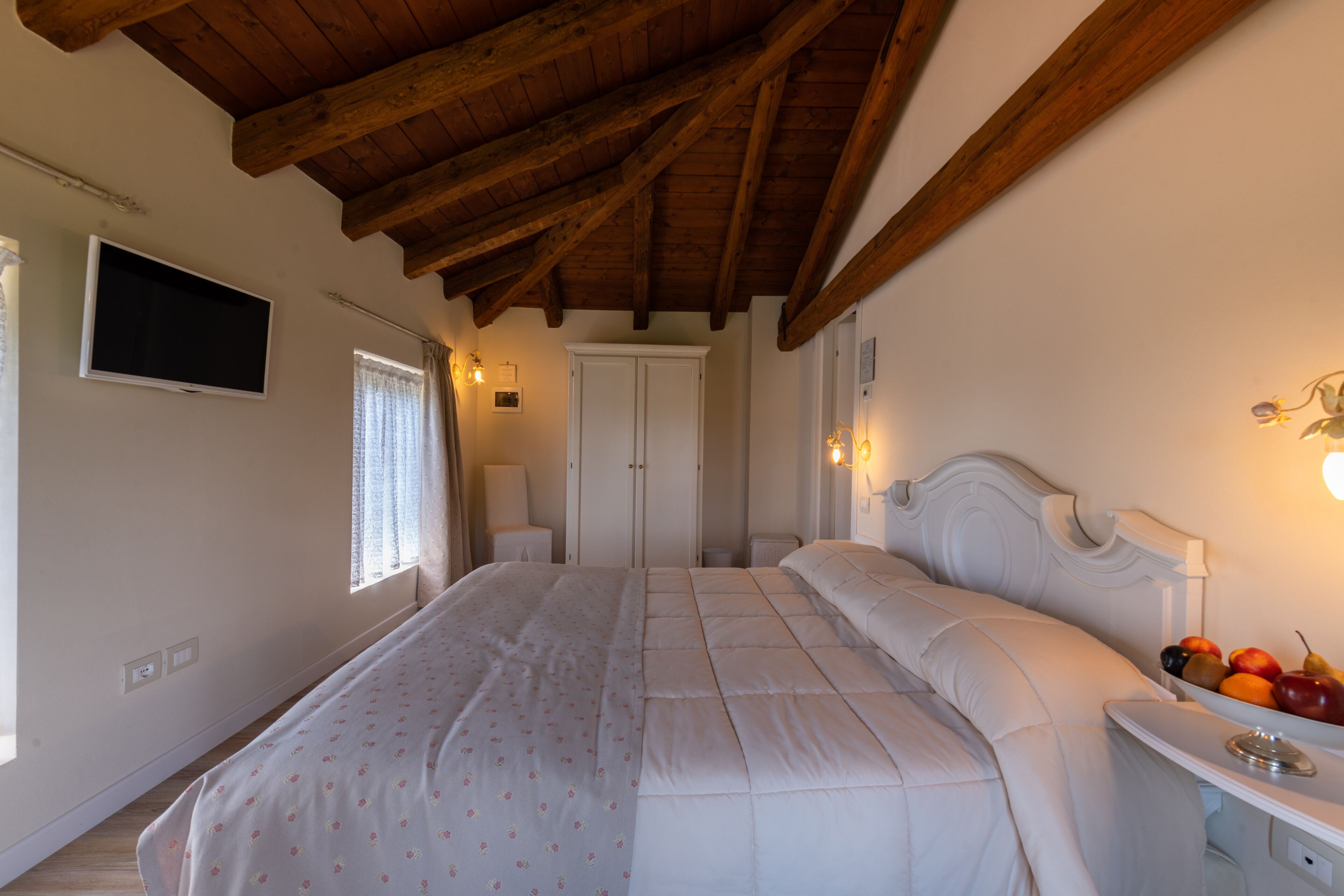 Une chambre avec un grand lit, des poutres apparentes en bois au plafond, un téléviseur fixé au mur et une table de chevet sur laquelle se trouvent des fruits.