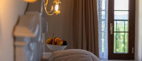 Un dormitorio con una cama, una mesita de noche con frutero, una ventana con cortinas y una lámpara de pared.