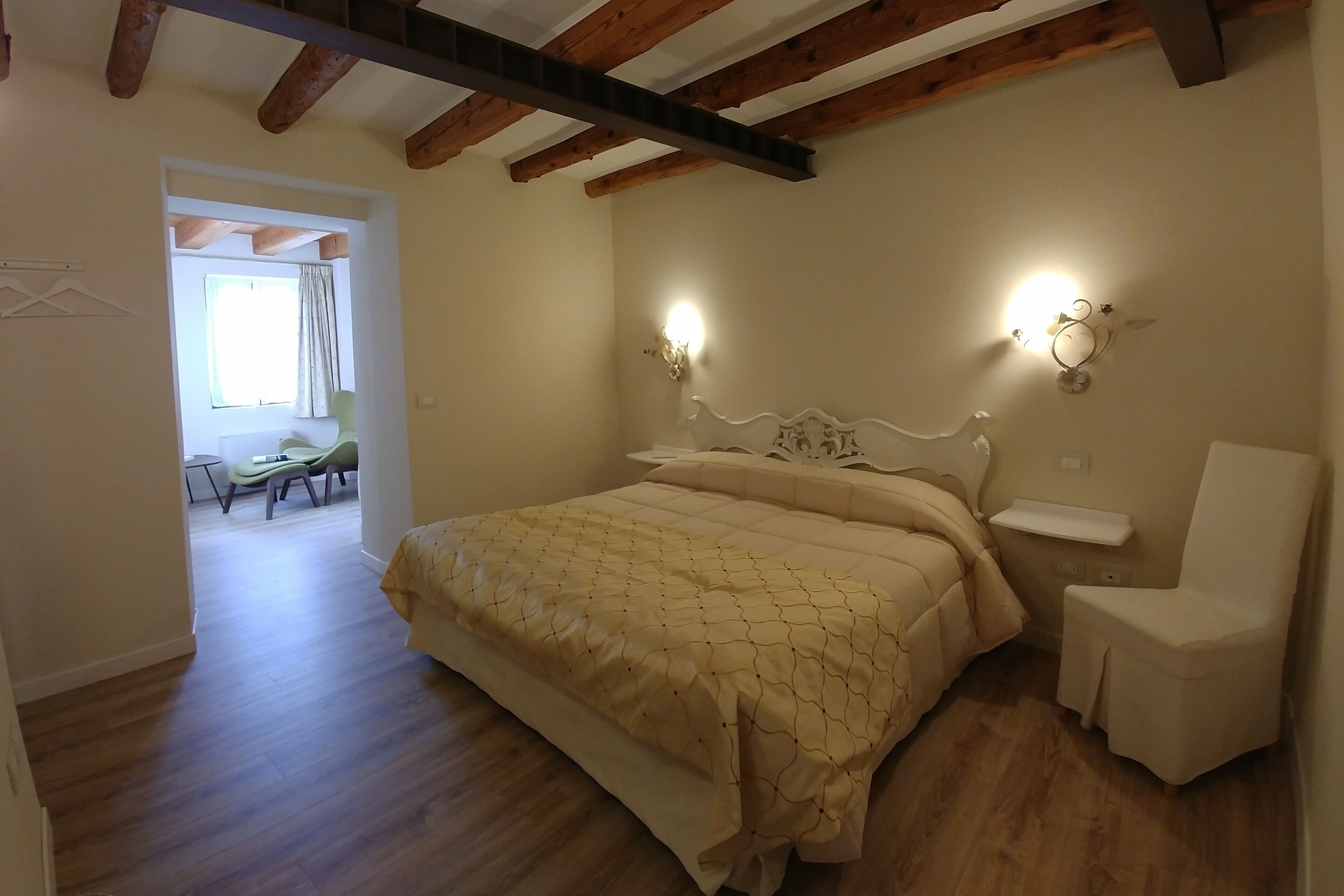 Un dormitorio con cama, mesitas de noche y una silla. Hay una ventana con cortinas y una puerta que da a otra habitación.