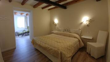 Un dormitorio con cama, mesitas de noche y una silla. Hay una ventana con cortinas y una puerta que da a otra habitación.