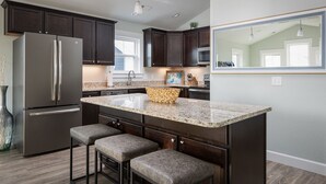 A modern kitchen with dark cabinets, a stainless steel refrigerator, granite countertops, and a large island with stools. - Dog Friendly - Sound View Home on private harbor. Amazing Sunsets-Fishing-Dock (Salvo)