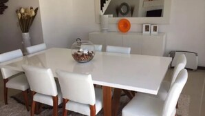 Un comedor con una mesa y sillas blancas, un cuenco de vidrio sobre la mesa, y una escalera al fondo.