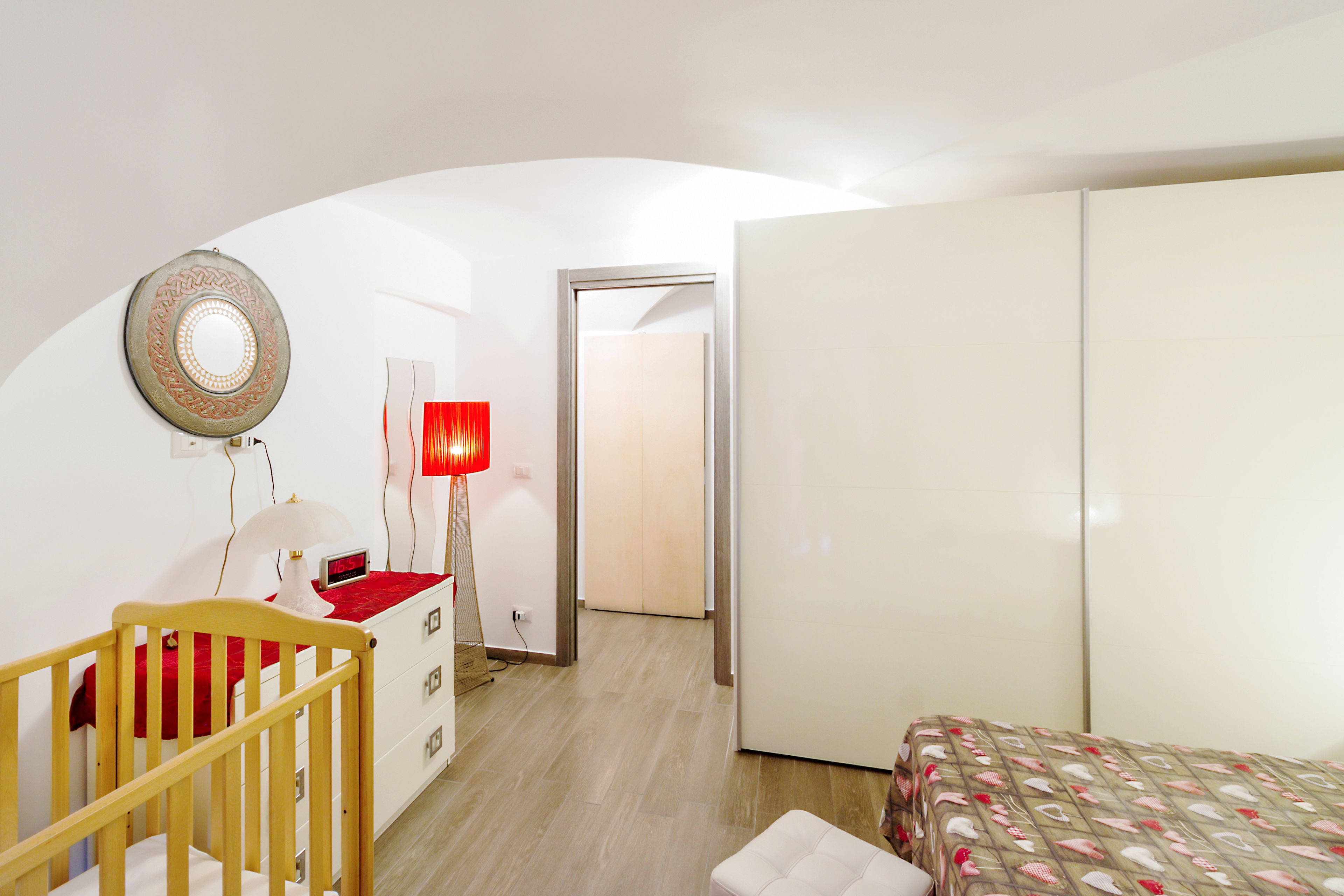 Un dormitorio con una cuna, un armario, una mesita de noche con lámpara y una colcha estampada.
