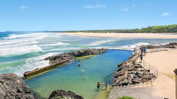 Zona de baño costera con agua cristalina, bordes rocosos y una playa de arena al fondo.