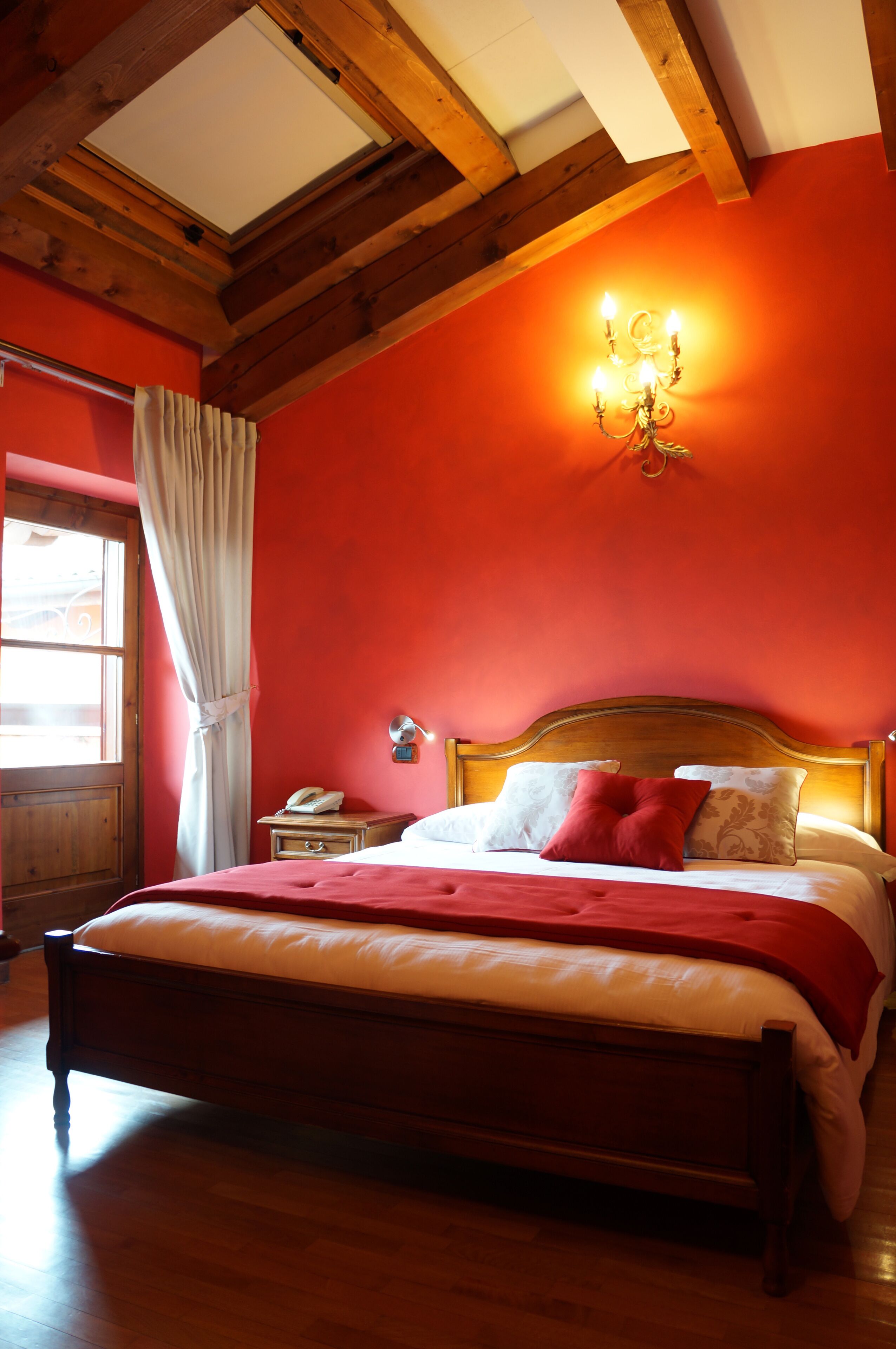 Un dormitorio con cama de madera, paredes rojas, techo de madera, mesita de noche con lámpara y ventana con cortinas.