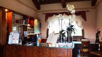 A reception area with a wooden desk, a bookshelf, a vase with flowers, and a window with curtains.