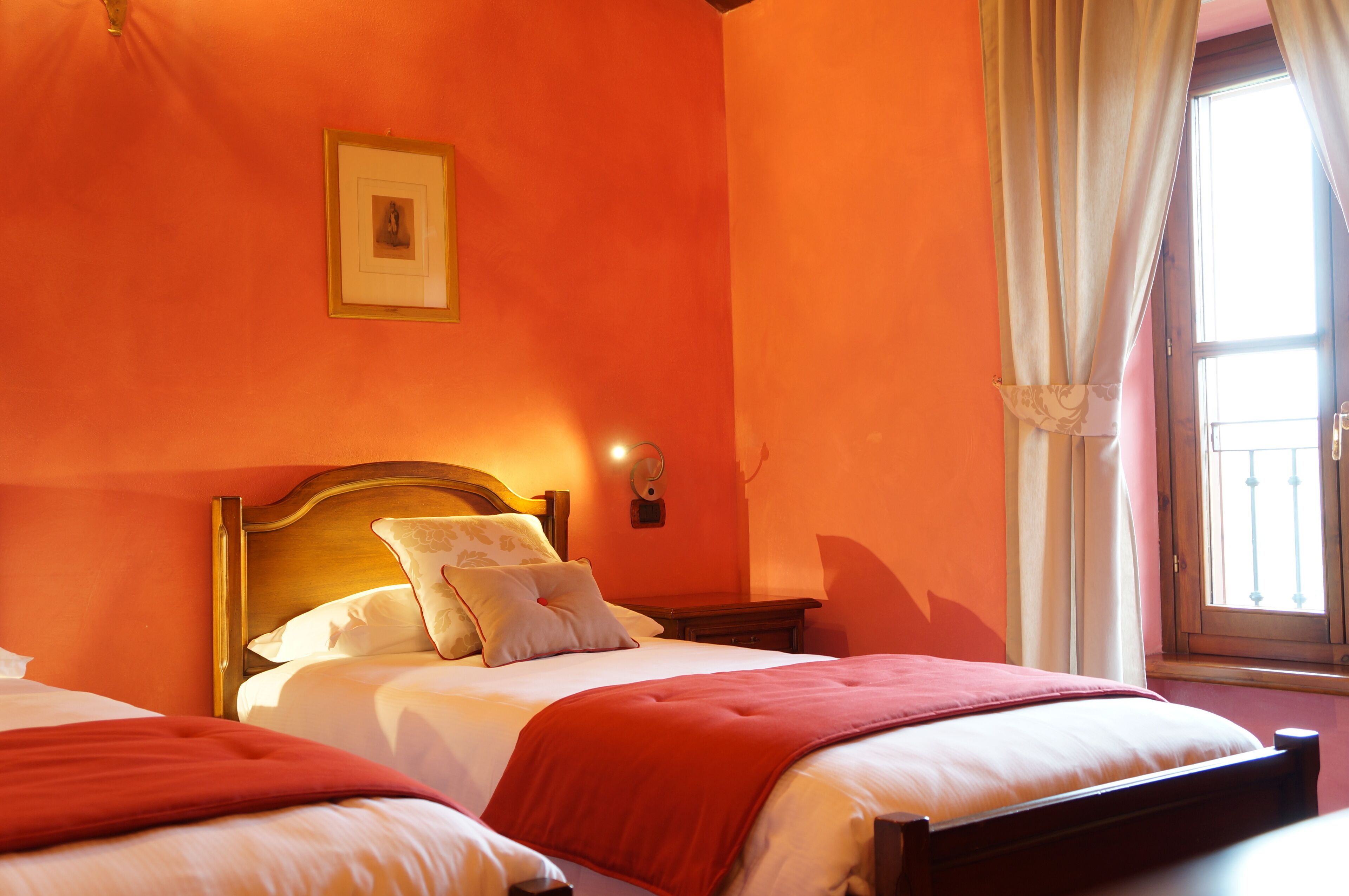 A hotel room with two beds, a wooden headboard, a nightstand, a wall-mounted light, and a window with curtains.