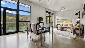 Ein großzügiger Wohnbereich mit Esstisch, Stühlen, einer Couch und einem Couchtisch. Große Fenster bieten einen Blick ins Freie.