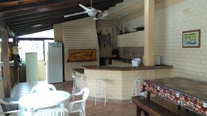 Cozinha com geladeira branca, pia e fogão. Há cadeiras brancas e uma mesa na área de refeições.