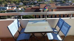 Een balkon met een tafel en blauwe stoelen, met uitzicht op een woongebied met huizen en een klokkentoren.