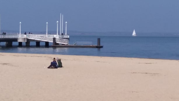 Een strand met een steiger die het water in reikt, een zeilboot in de verte en een paar mensen op het zand.