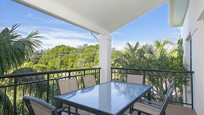 Un balcón con una mesa y sillas de vidrio, rodeado por una baranda blanca, con vistas a un paisaje verde y exuberante.