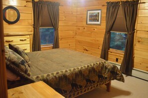 Una habitación de cabaña de madera con una cama, dos ventanas con cortinas, un espejo y un cuadro enmarcado en la pared.