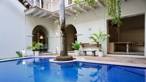 A swimming pool with a tree in the center, surrounded by potted plants and a patio area with a table and chairs.