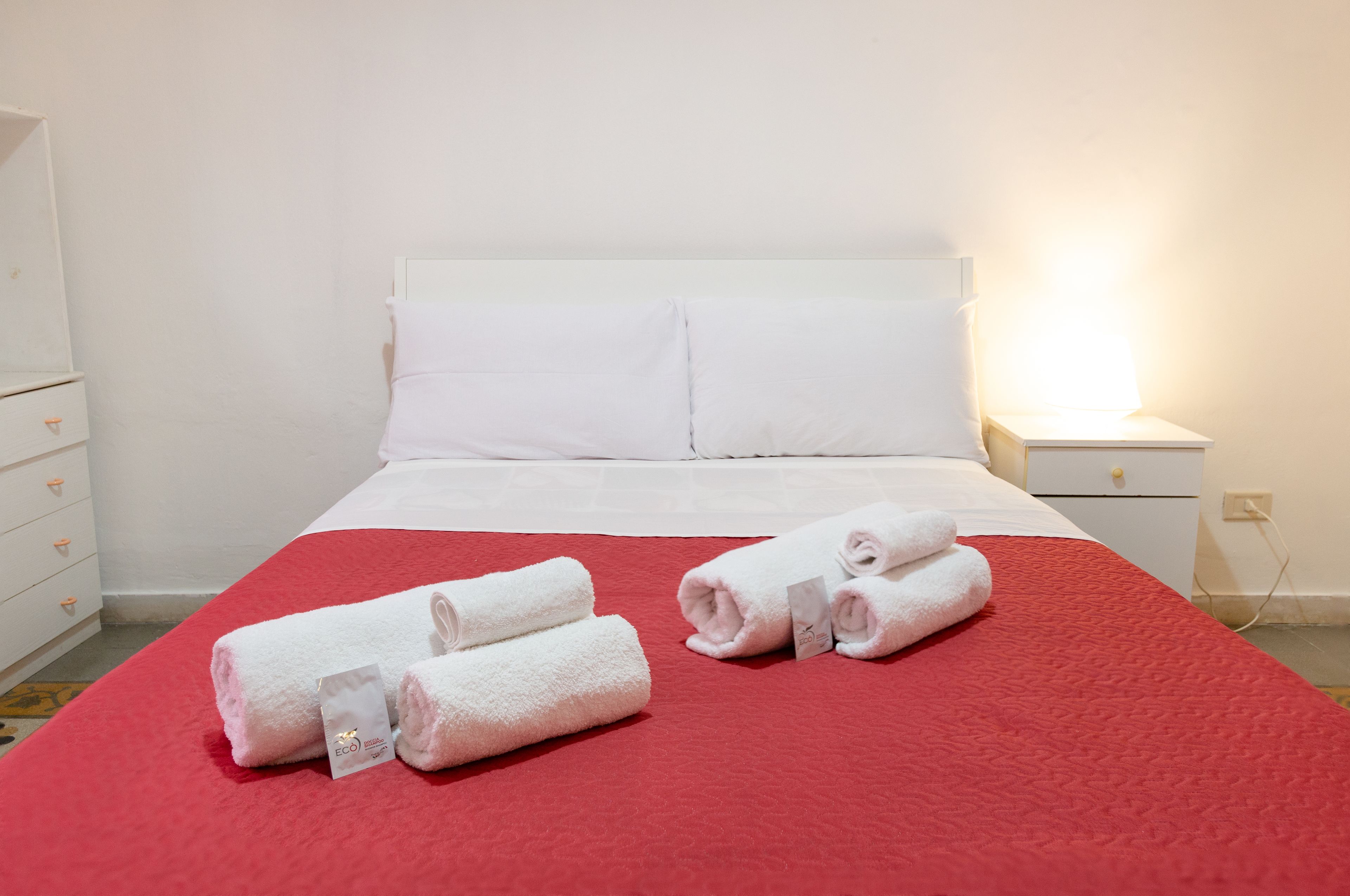 Habitació d'hotel amb un llit, almoines blanques, un llençol vermell i dues tovelloles blanques amb etiquetes sobre el llit.
