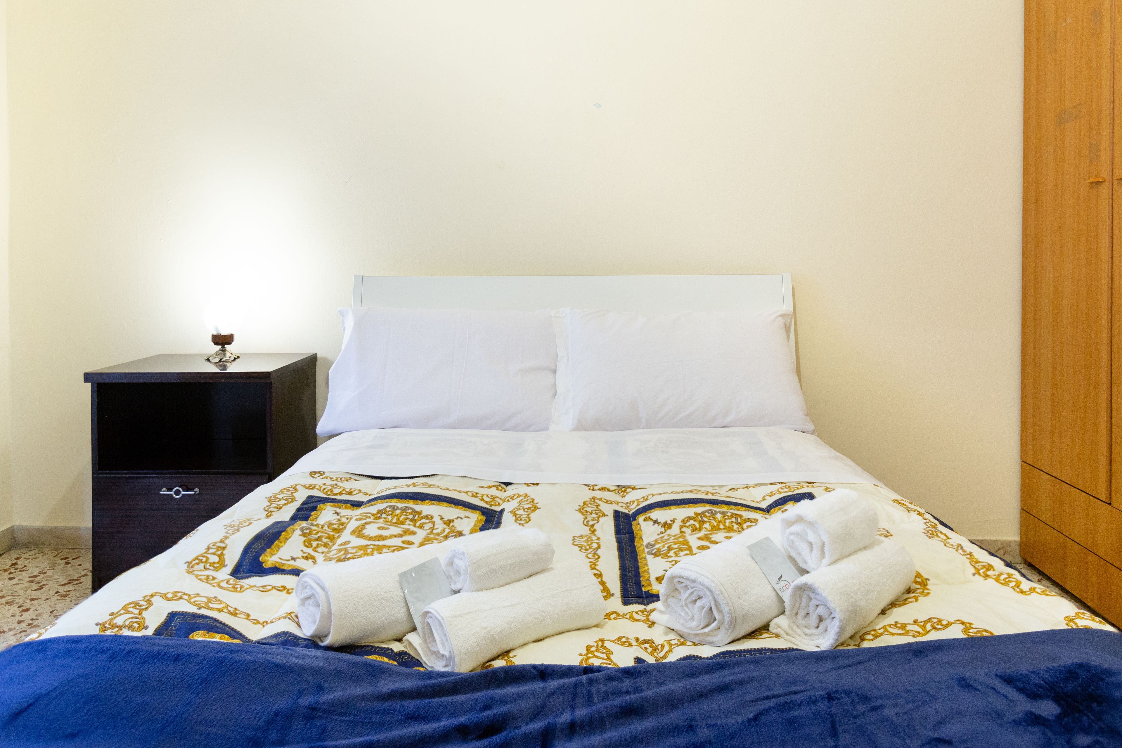 A neatly made bed with a patterned bedspread, a dark nightstand with a small item on it, and a wooden wardrobe.