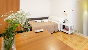 Ein Hotelzimmer mit Holzboden, einem Bett mit weißen Leinen, einem Nachttisch mit Fernbedienung, einem kleinen weißen Beistelltisch und einer Vase mit Blumen auf der linken Seite.
