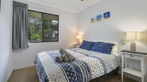 A bedroom with a bed, two bedside tables, a window with curtains, and wall art. - Ocean Sands 5 is a fully furnished two bedroom unit located on the first floor, next to the beach. (Sawtell)