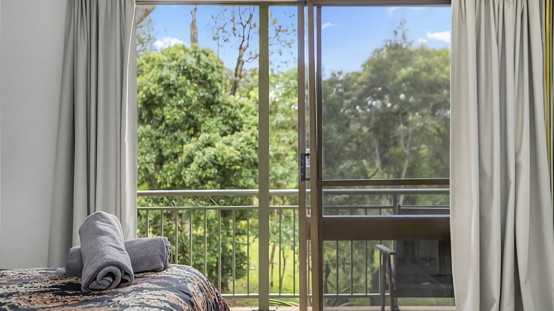 A room with a bed, a window with a view of greenery, and a balcony with a railing.