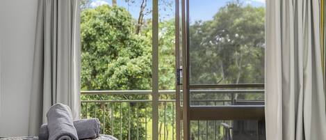A room with a bed, a window with a view of greenery, and a balcony with a railing.