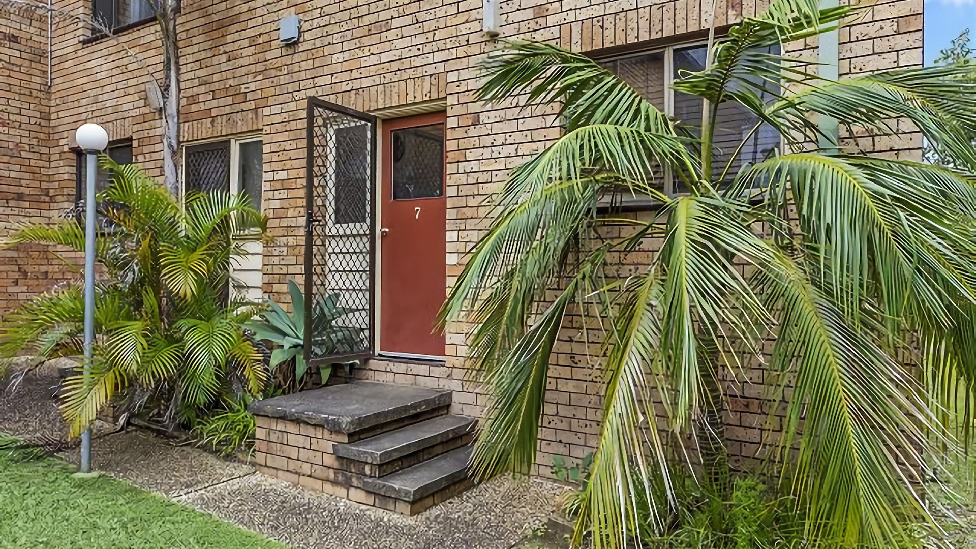 A brick building with a red door, a small front porch, and a number '7' on the door.