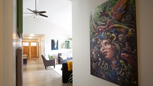 Un couloir avec un ventilateur de plafond, un tableau représentant une femme entourée d’animaux, et un espace de vie comprenant un canapé et une table basse.