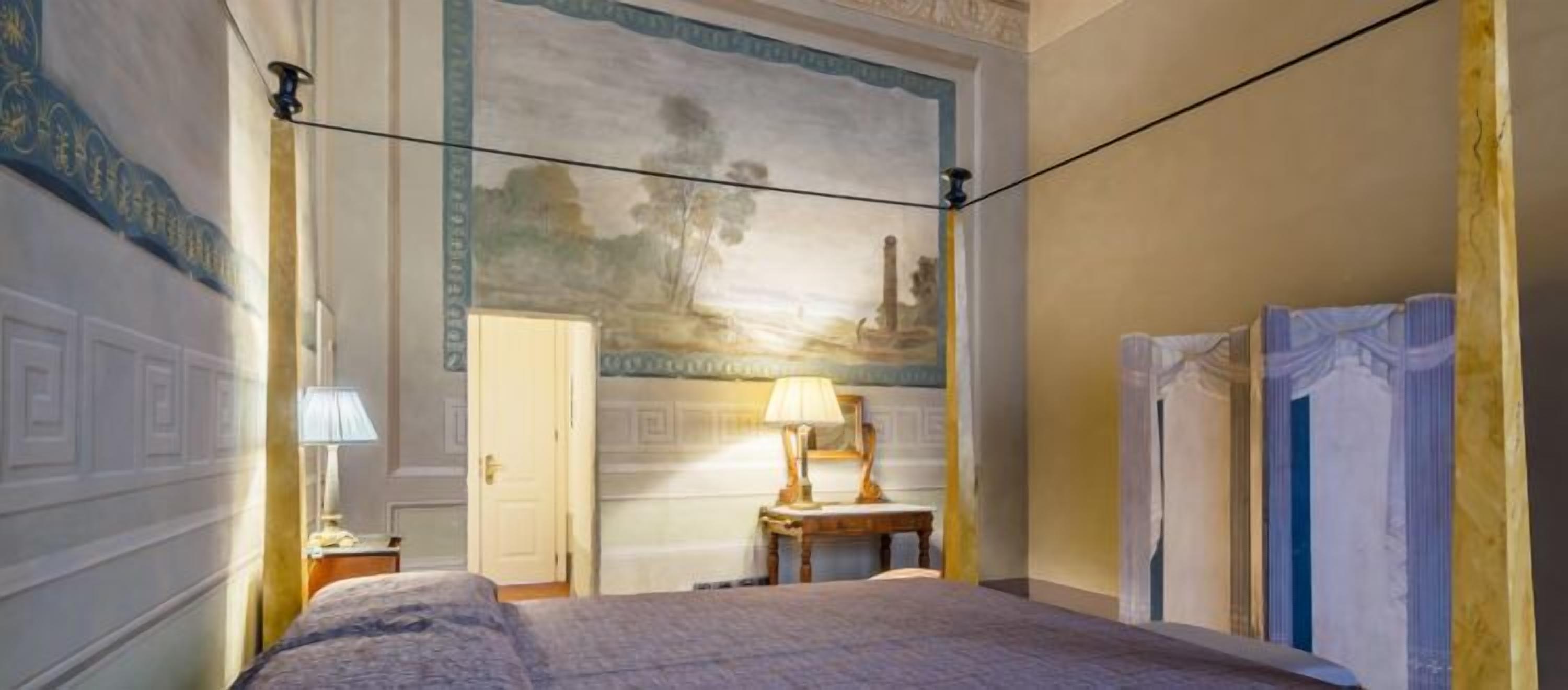 Ein Hotelzimmer mit Bett, Nachttisch, Lampe, Tür und einer Wand mit Wandgemälde.