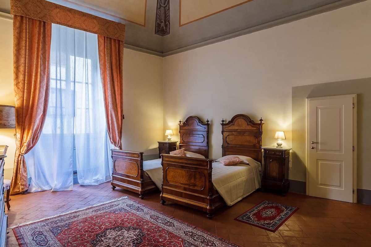 A traditional bedroom with a double bed, two bedside tables, a nightstand, a large window with curtains, and a patterned rug.