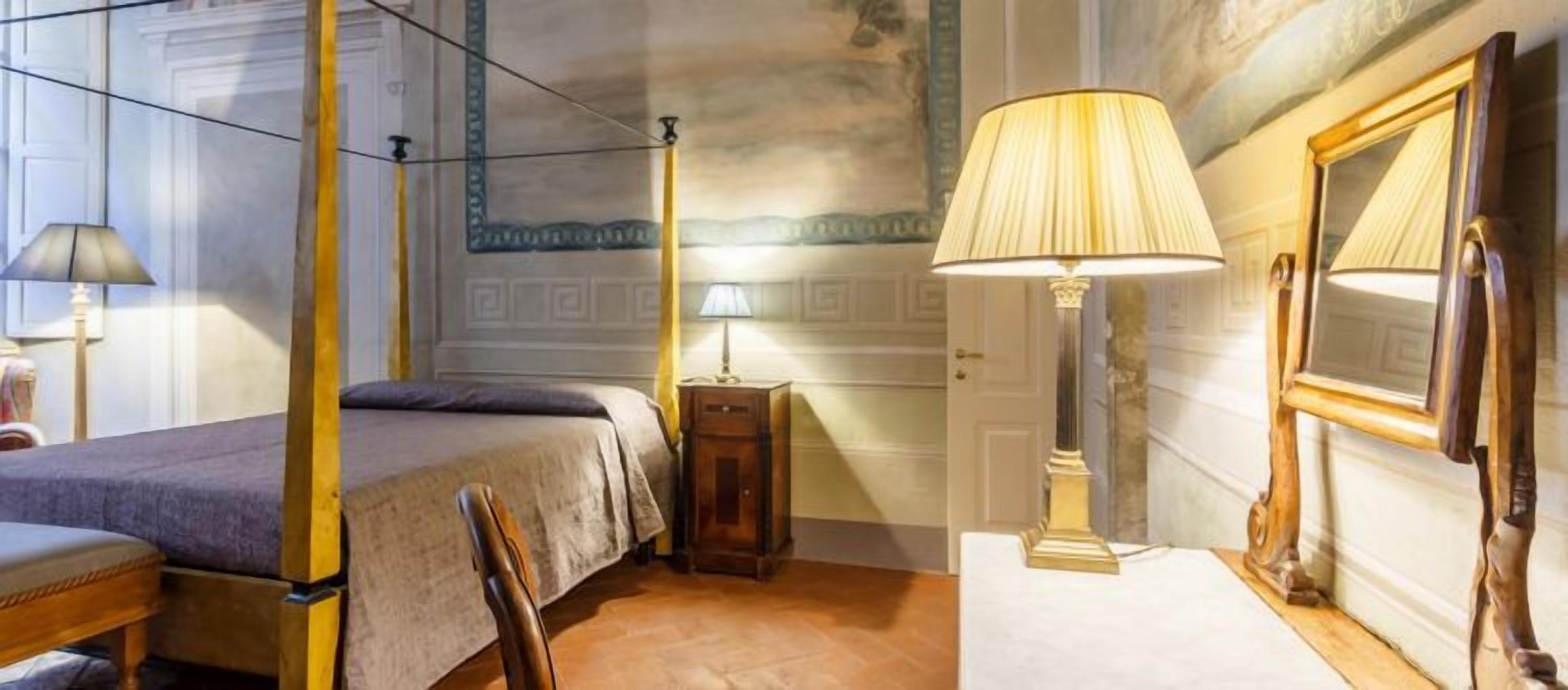 A four-poster bed with a wooden headboard, a bedside table with a lamp, a chair, and a mirror on the wall.