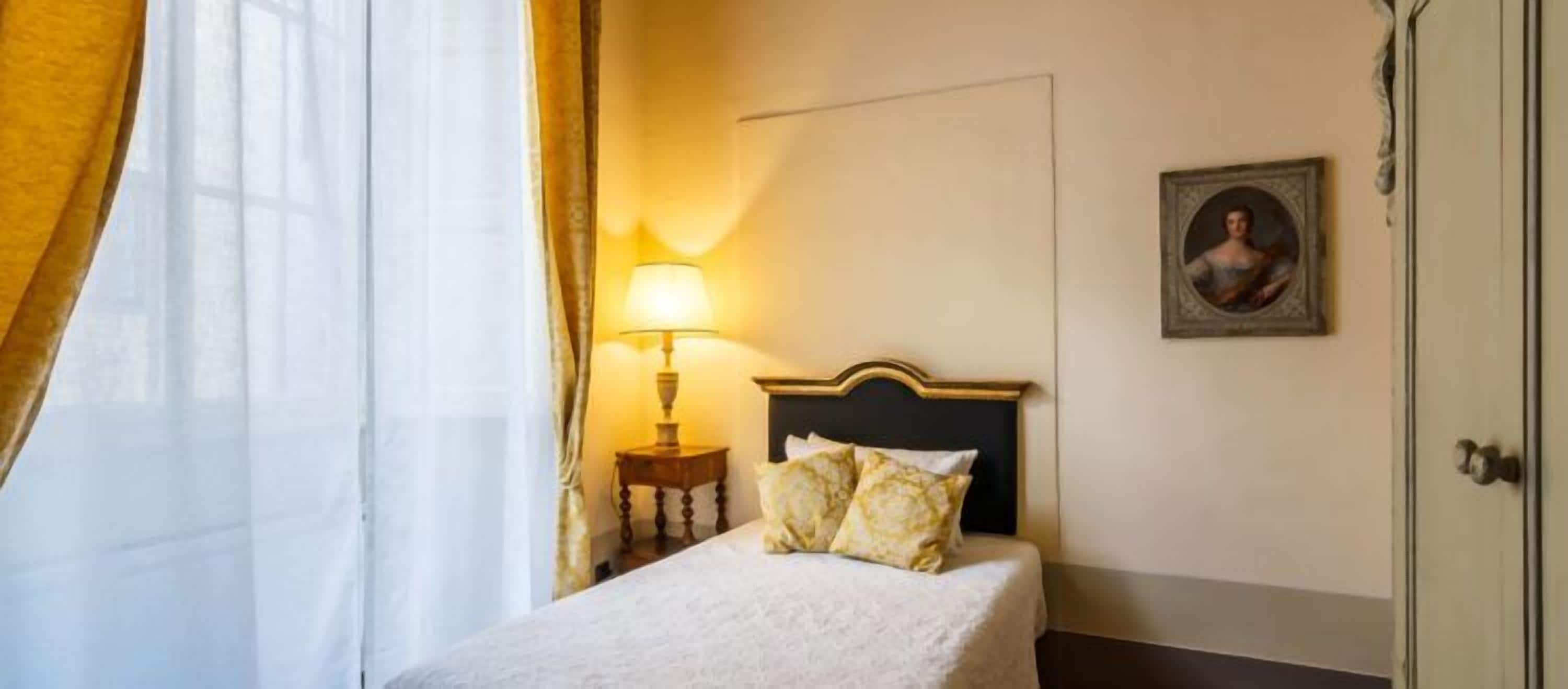 Una habitación de hotel con una cama, una mesita de noche con lámpara, un cuadro en la pared y una puerta.