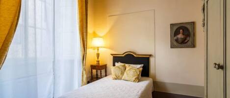 Una habitación de hotel con una cama, una mesita de noche con lámpara, un cuadro en la pared y una puerta.