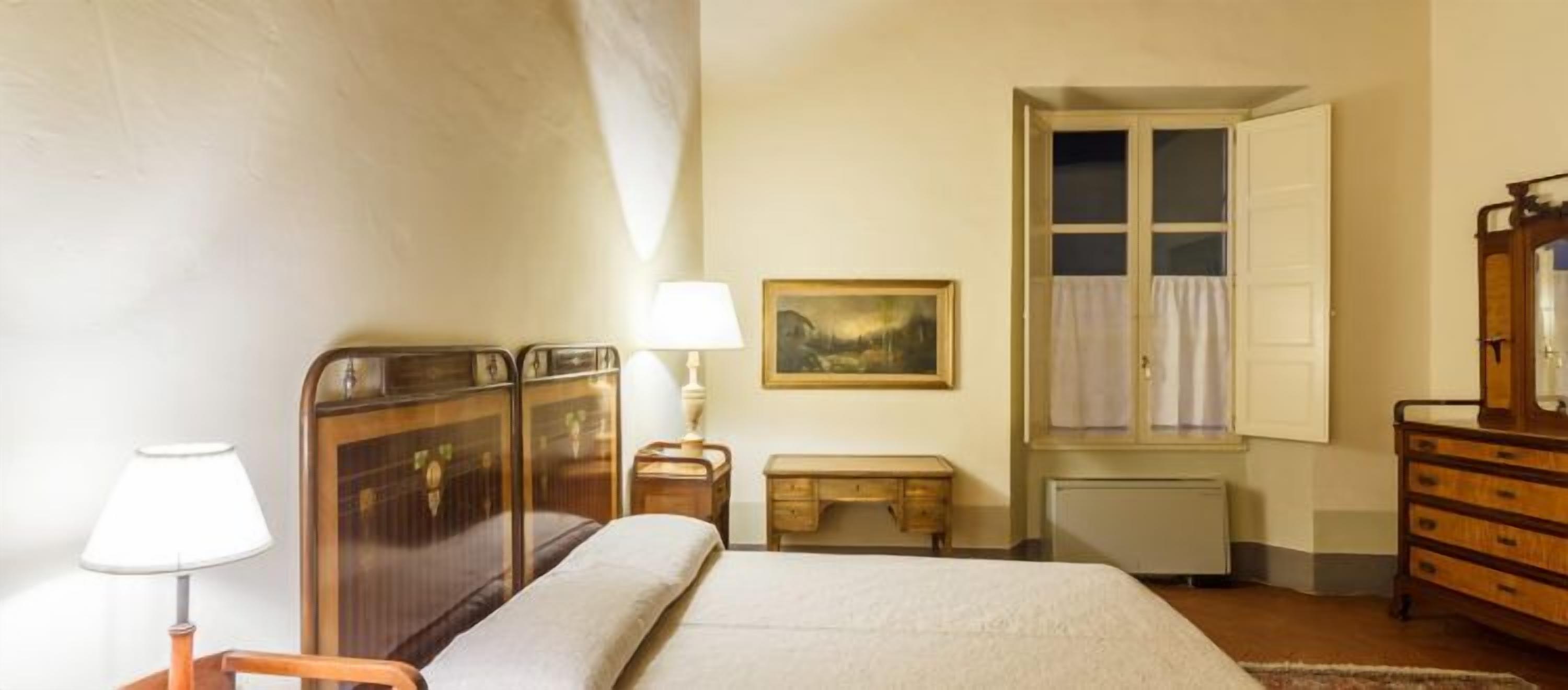 A bedroom with a bed, a desk, a lamp, a dresser, a window with curtains, and a painting on the wall.