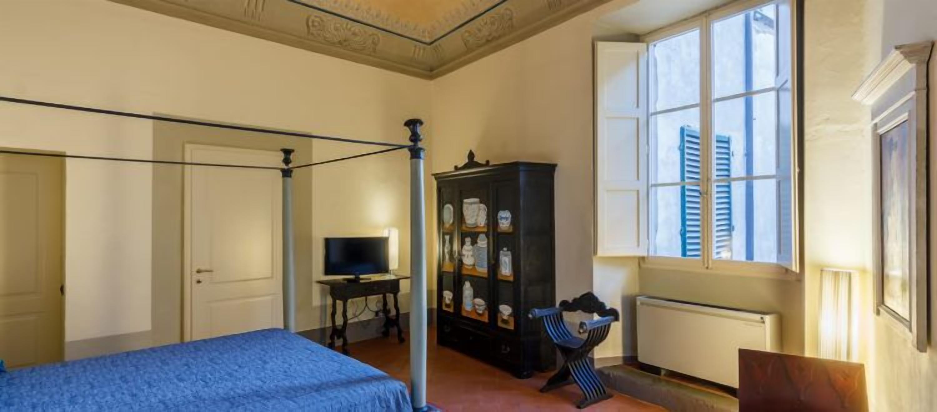 A bedroom with a four-poster bed, a television on a side table, a cabinet with decorative items, and a window with shutters.