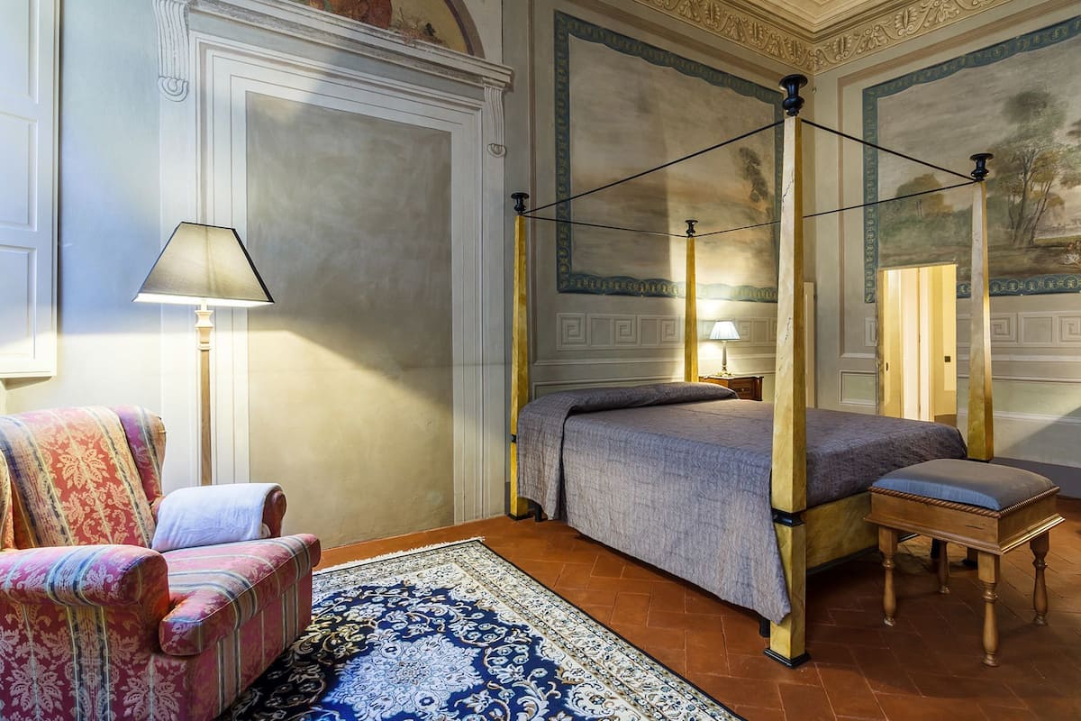 A four-poster bed with a canopy, a patterned armchair, a small wooden table, and a floor lamp in a room with ornate wall decorations and a tiled floor.