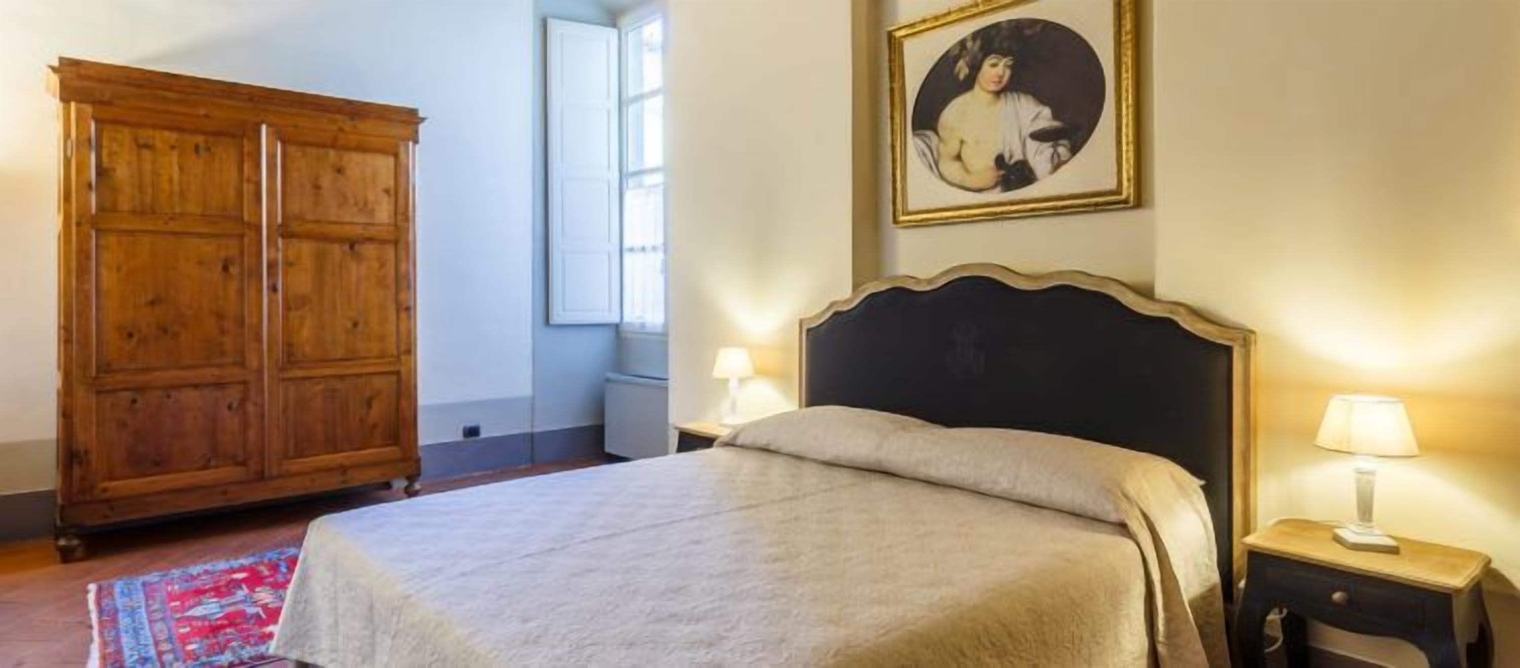 A bedroom with a large wooden wardrobe, a bed with a dark headboard, two bedside lamps, and a framed picture on the wall.