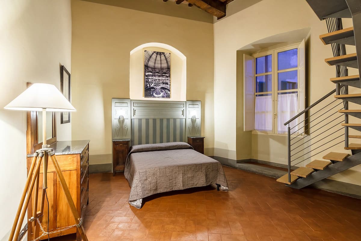 A bedroom with a bed, a lamp, a dresser, a window, and a staircase.