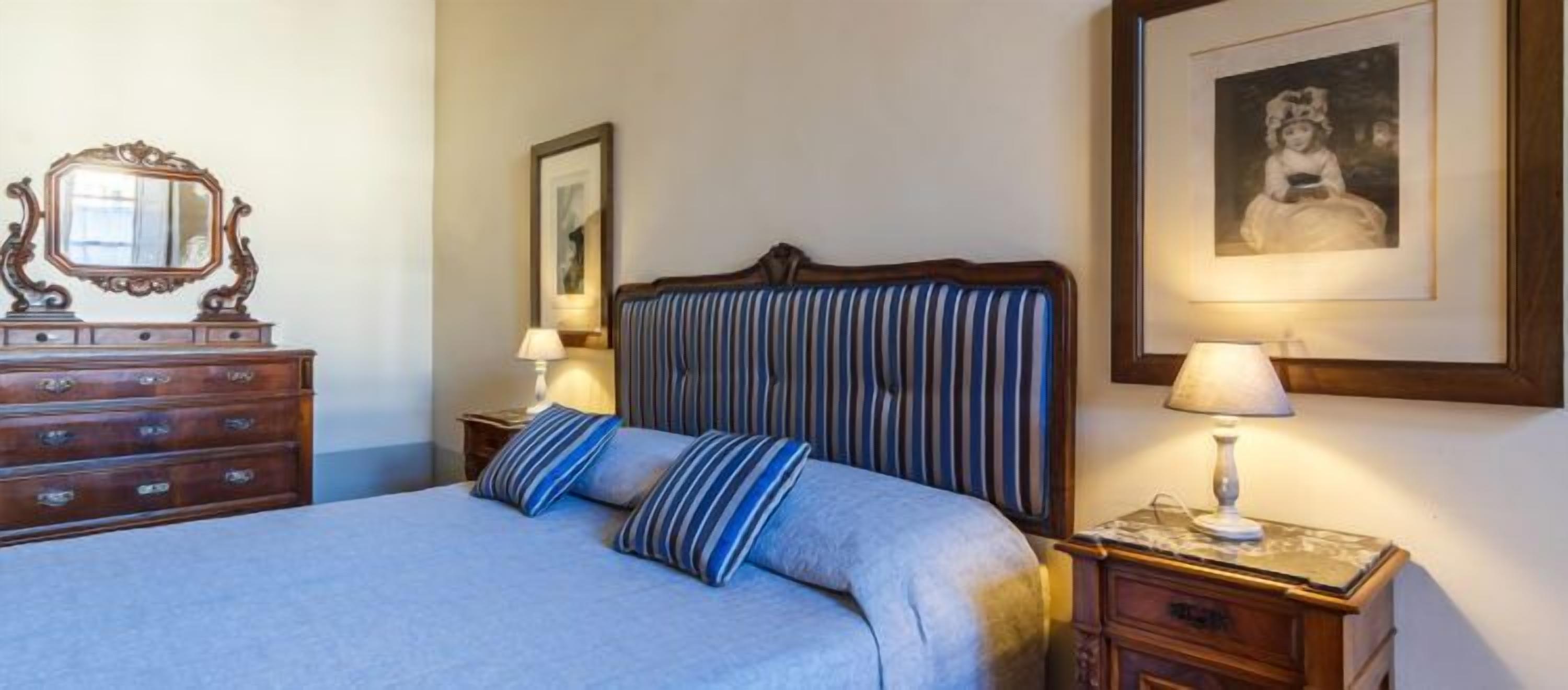 Een opgemaakt bed met gestreepte kussens, een houten dressoir met lades en een ingelijst kunstwerk aan de muur.