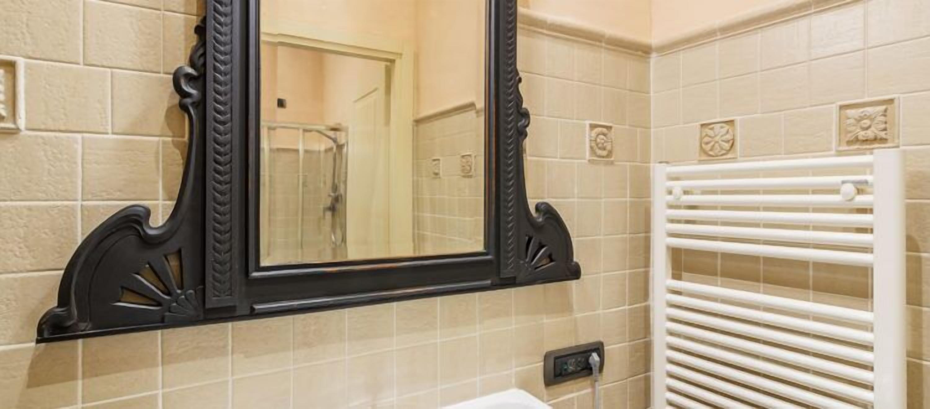 Un baño con un espejo grande, paredes cubiertas de baldosas color beige, un perchero blanco y una zona de ducha visible.