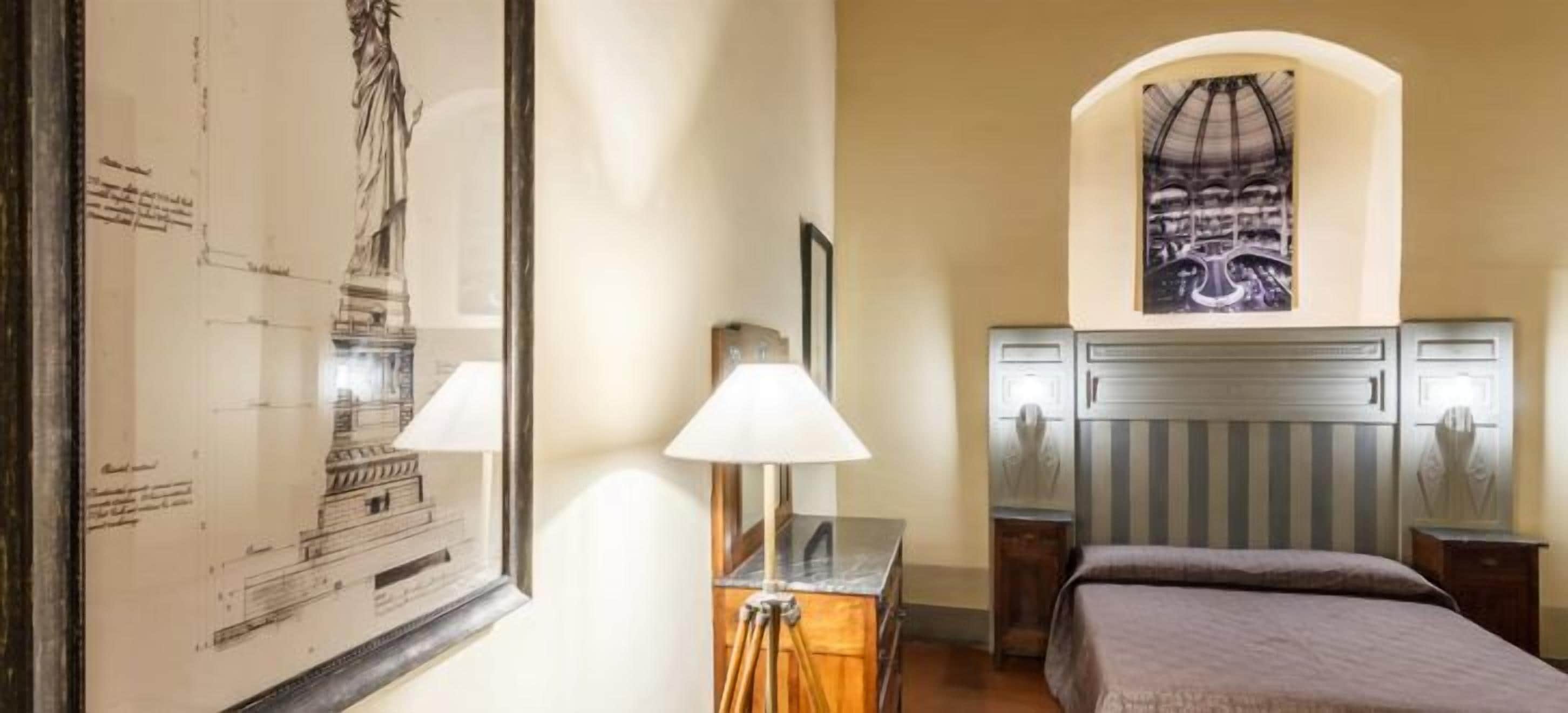 A hotel room with a bed, a desk, a lamp, a framed architectural drawing, and a painting on the wall.