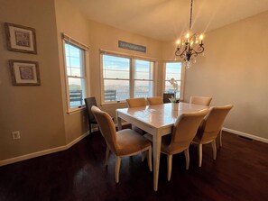 A dining room with a white table and six chairs, large windows with a view, a chandelier, and framed pictures on the wall.
