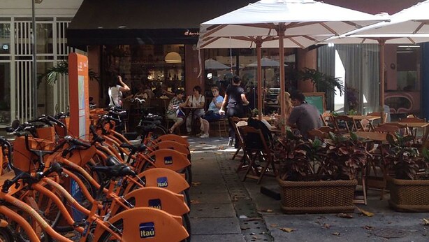 A row of orange bicycles parked outside a building with outdoor seating and umbrellas. - Excelente Apartamento Em Copacabana (Rio de Janeiro)