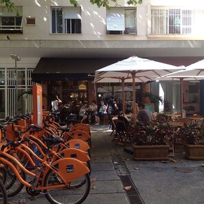 A row of orange bicycles parked outside a building with outdoor seating and umbrellas. - Excelente Apartamento Em Copacabana (Rio de Janeiro)