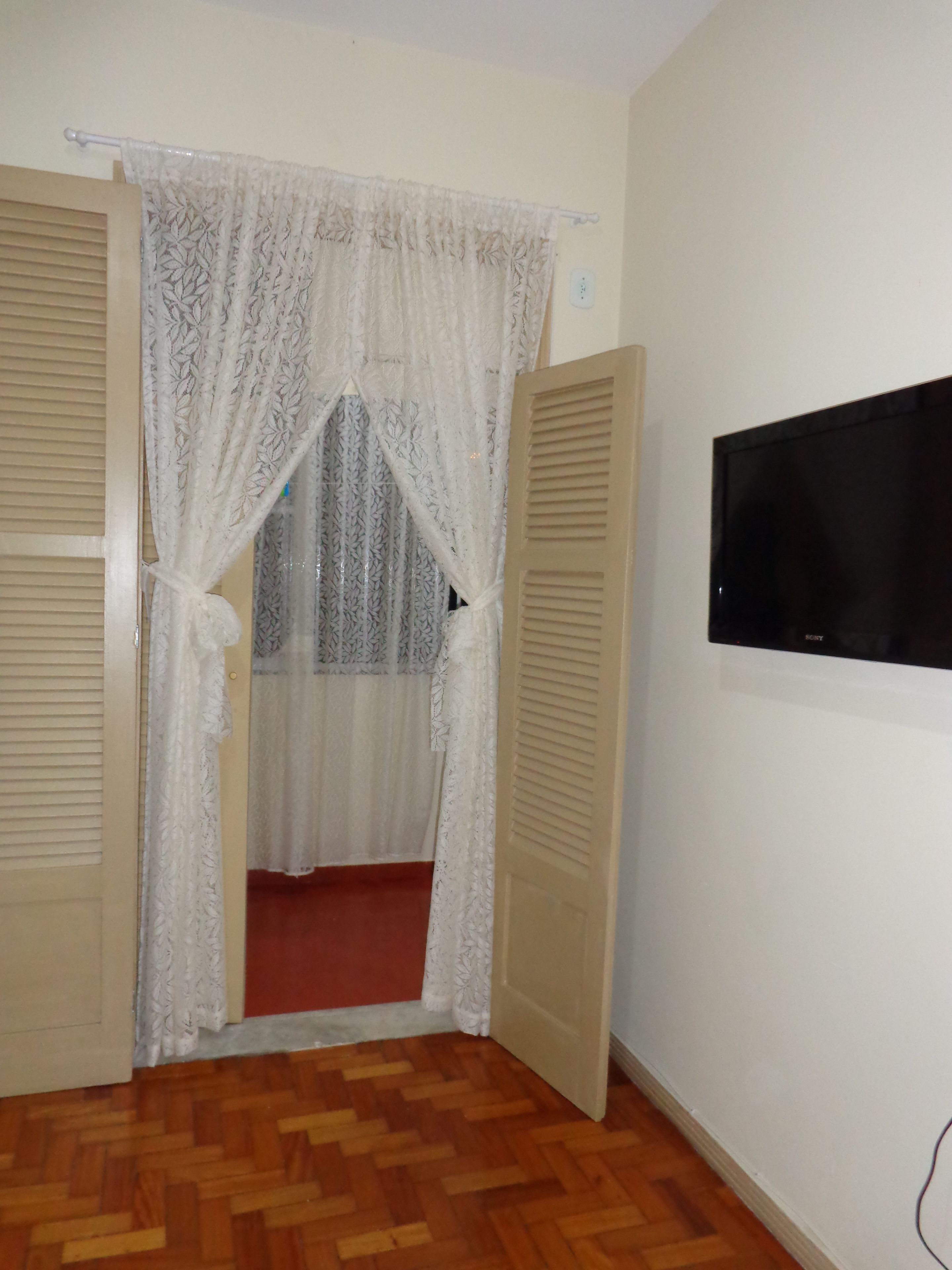An open door with a white lace curtain, a wooden floor, and a wall-mounted TV.