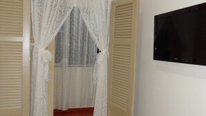 An open door with a white lace curtain, a wooden floor, and a wall-mounted TV. - Excelente Apartamento Em Copacabana (Rio de Janeiro)