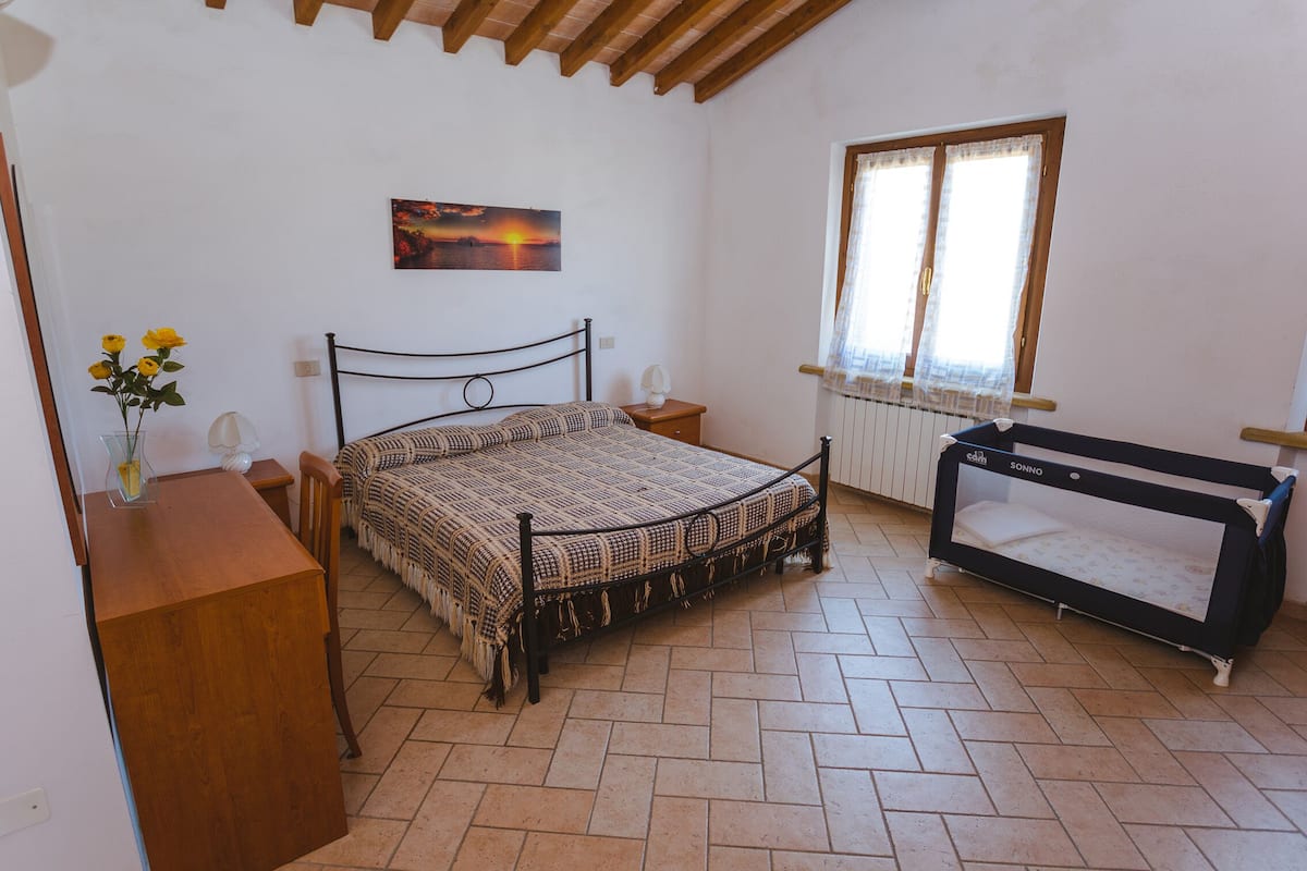 A bedroom with a bed, a wooden dresser, a nightstand, a baby crib, a window with curtains, and a painting on the wall.