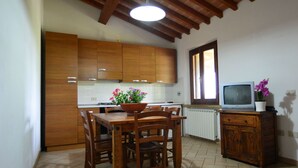 A kitchen with wooden cabinets, a dining table with chairs, a flower pot, a television on a cabinet, and a window with natural light. - Agriturismo Ferri di Ferri Donatella (Bibbona)