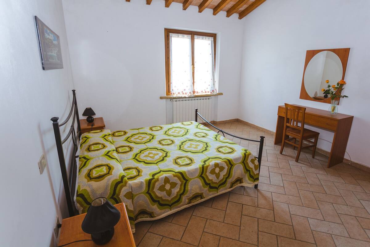 A bedroom with a bed, a wooden desk, a chair, a mirror, and a window with curtains.