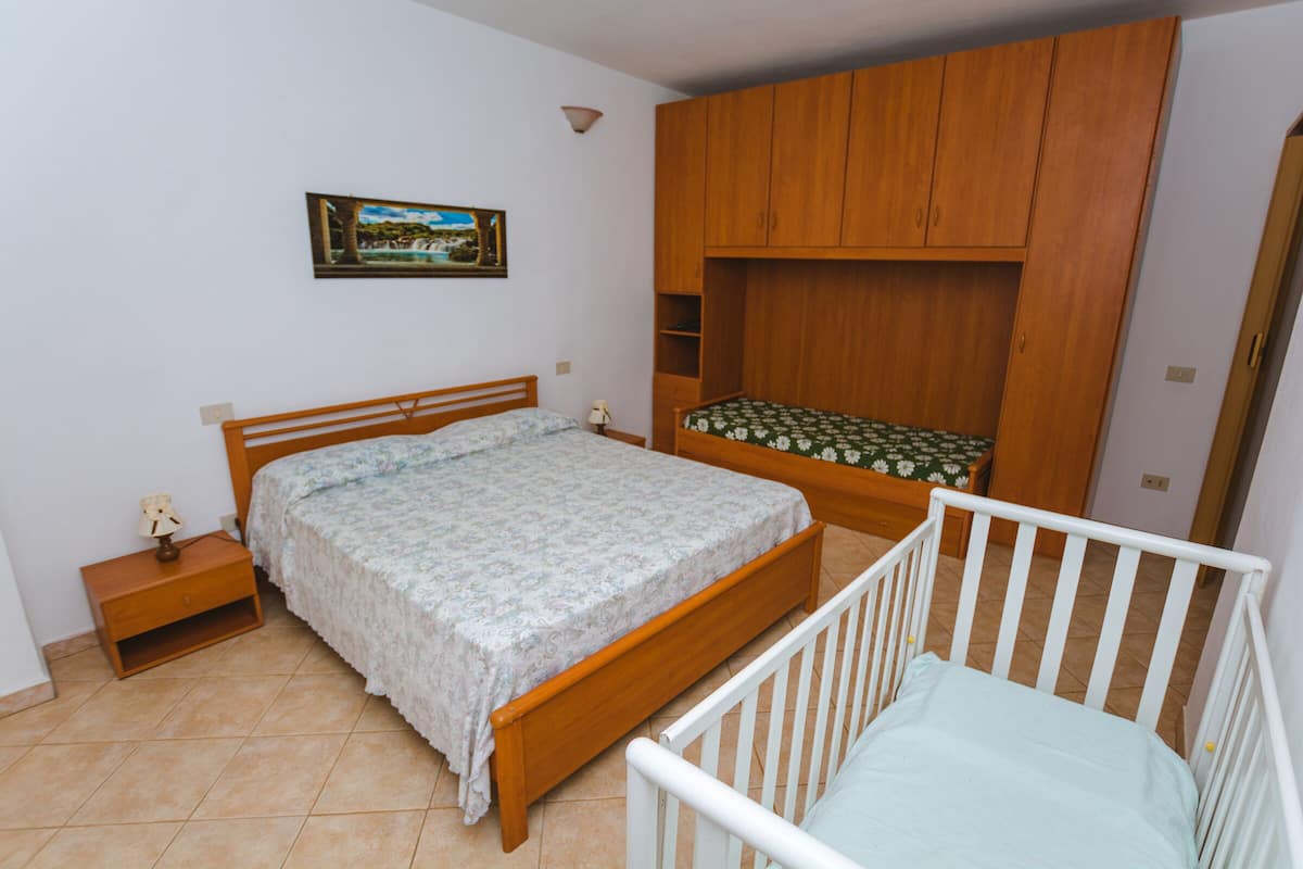 A bedroom with a wooden bed, a crib, a nightstand, a wall-mounted shelf, and a painting.